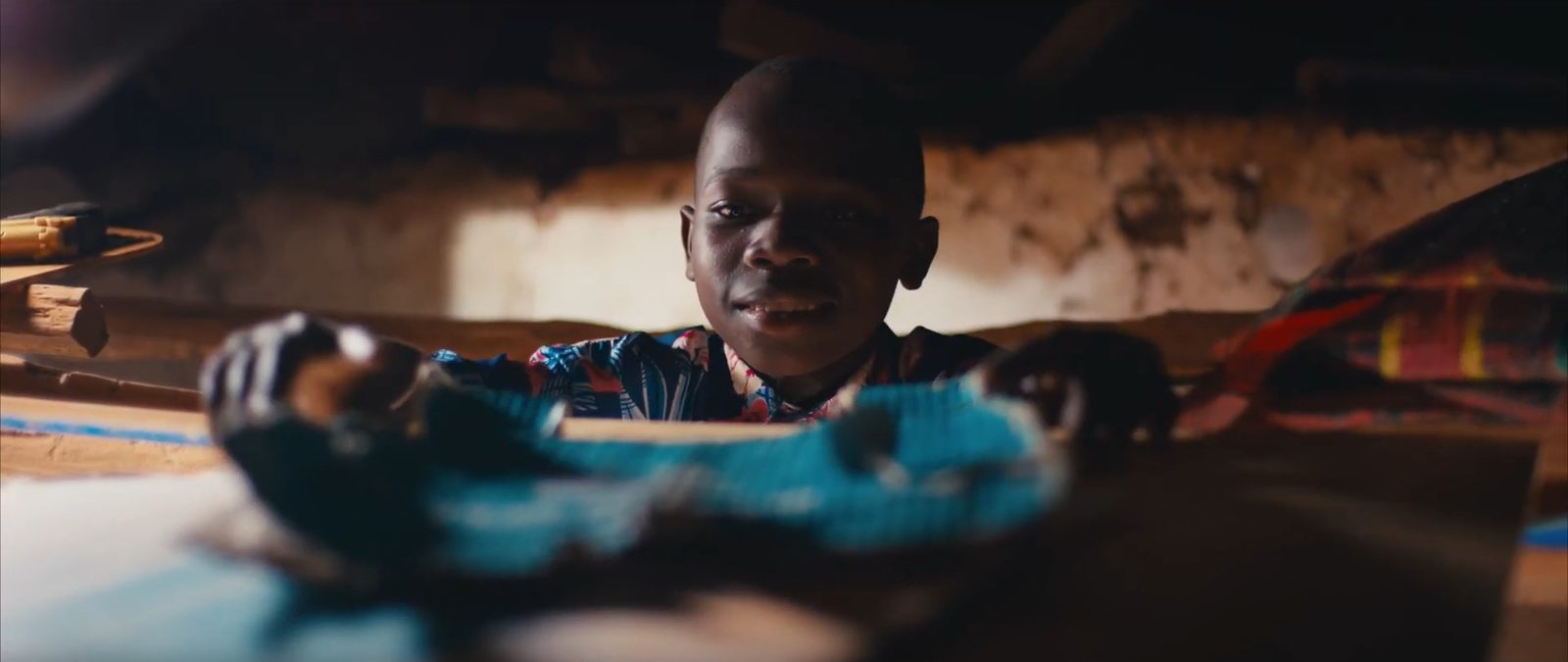 A smiling boy looks over a table in a hut