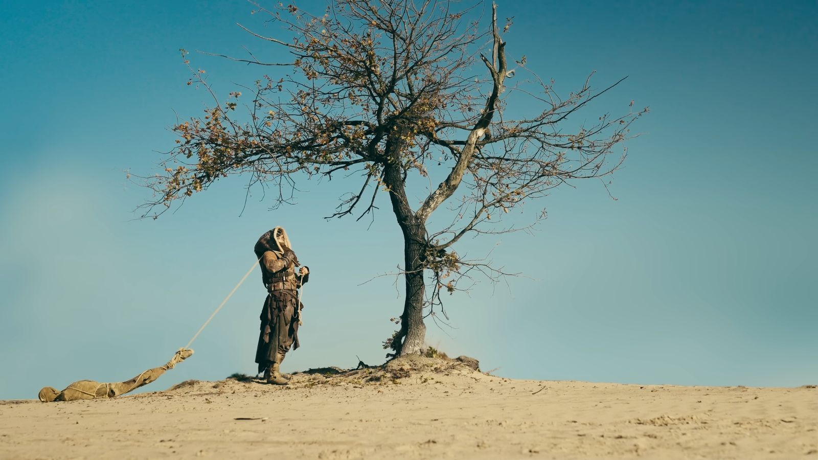 A hooded wanderer pulls a bound figure past a lone tree